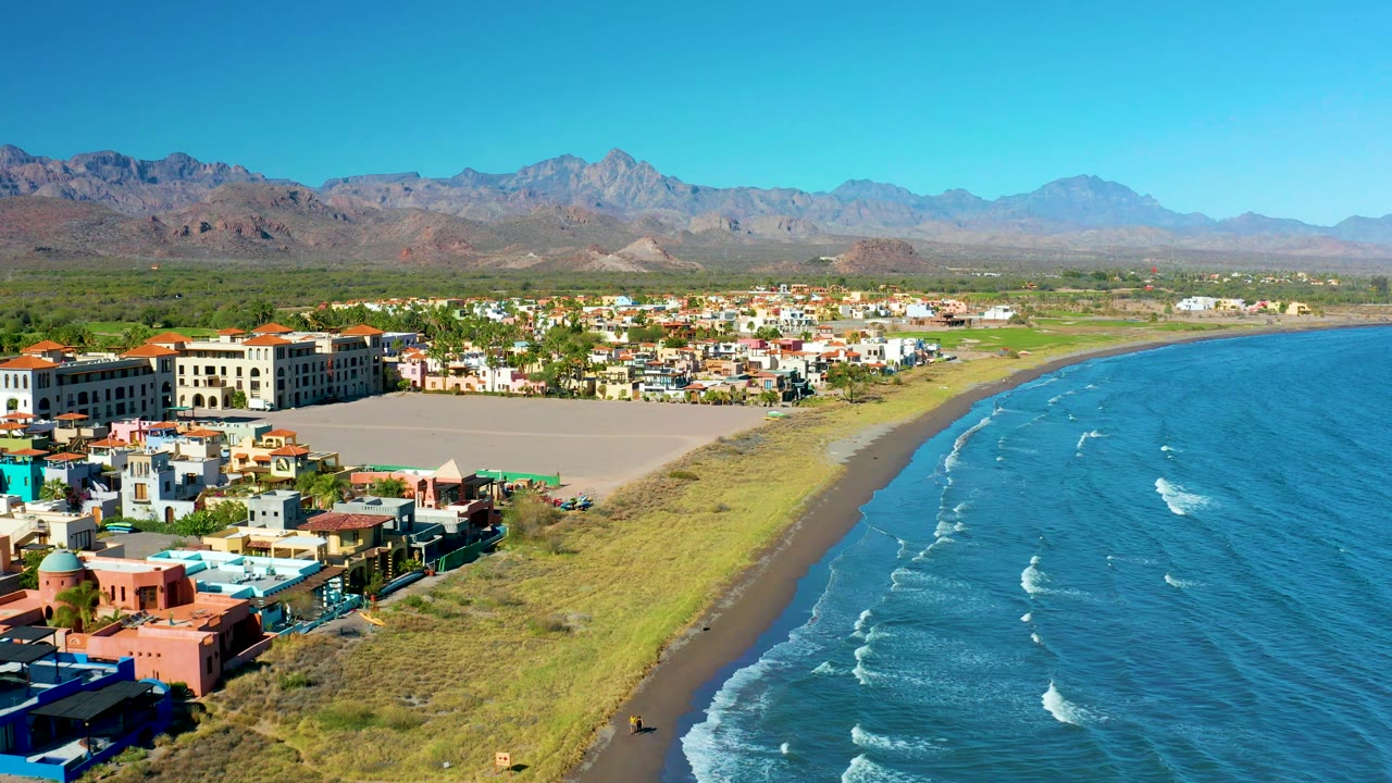 Wide aerial frame over Loreto, Baja California Sur showing colorful buildings along the coast, grassy dunes between town and water, the ocean filling the right side, distant mountains, and no visible people.