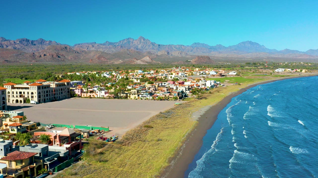 Aerial view over Loreto, Baja California Sur with a sandy beach, blue ocean waves, low buildings, distant mountains, clear sky, and no visible people.