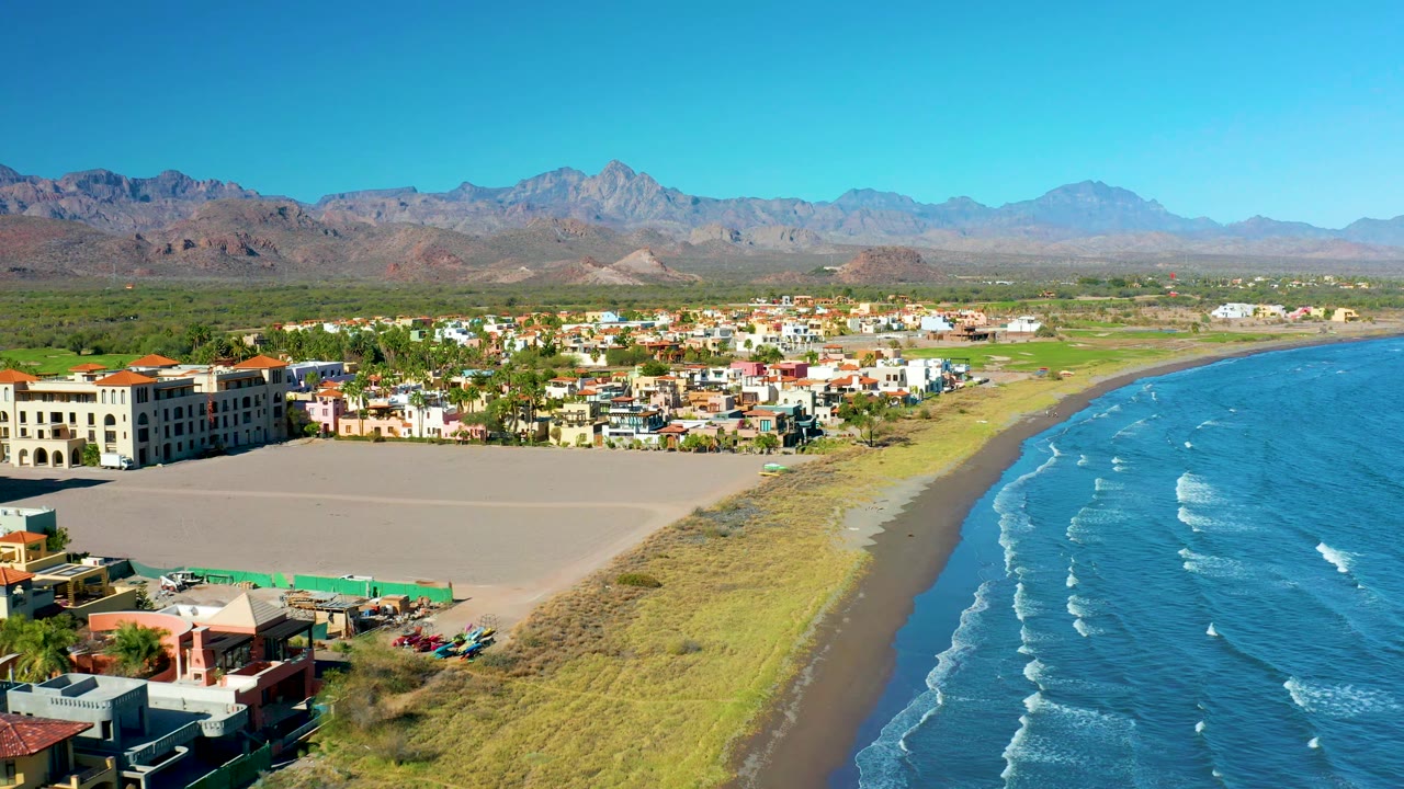 Aerial view of Loreto's curving beach with blue waves at right, compact town blocks near the shore, open sandy ground, mountains behind, and no visible people.