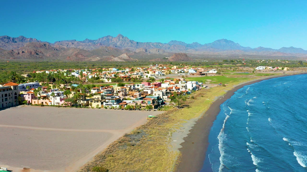 Aerial frame over Loreto, Baja California Sur, with a wide sandy beach at left, blue ocean at right, low coastal buildings in the middle, mountains on the horizon, and no visible people.