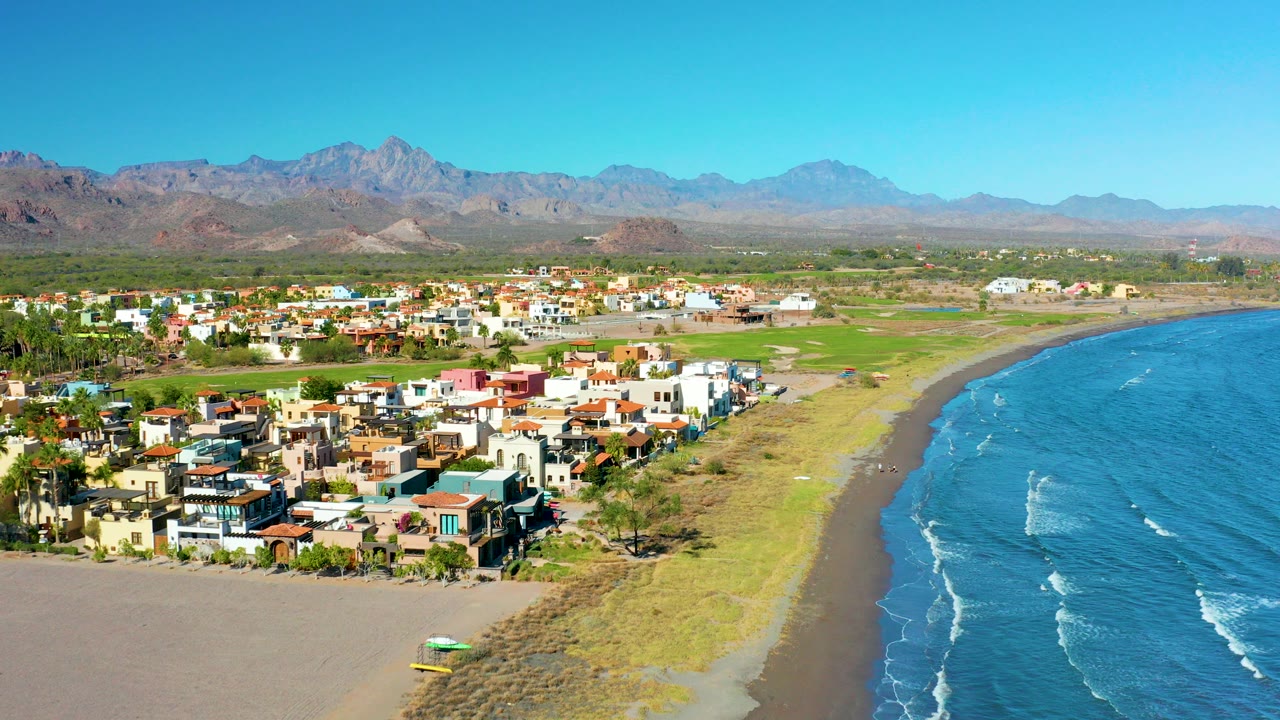 Aerial frame over Loreto, Baja California Sur with colorful homes at lower left, sandy beach and blue ocean at right, green land behind town, distant mountains, clear sky, and no visible people.