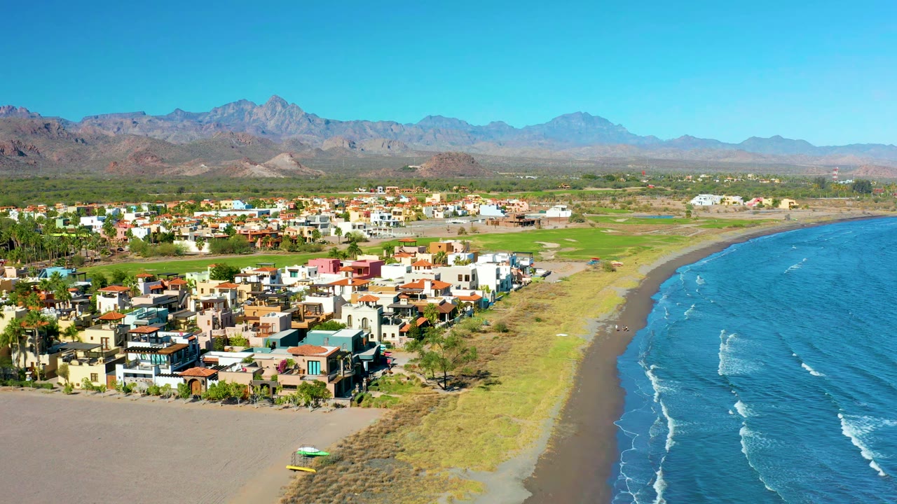 Aerial coastal town beside a curving beach with ocean waves and mountains
