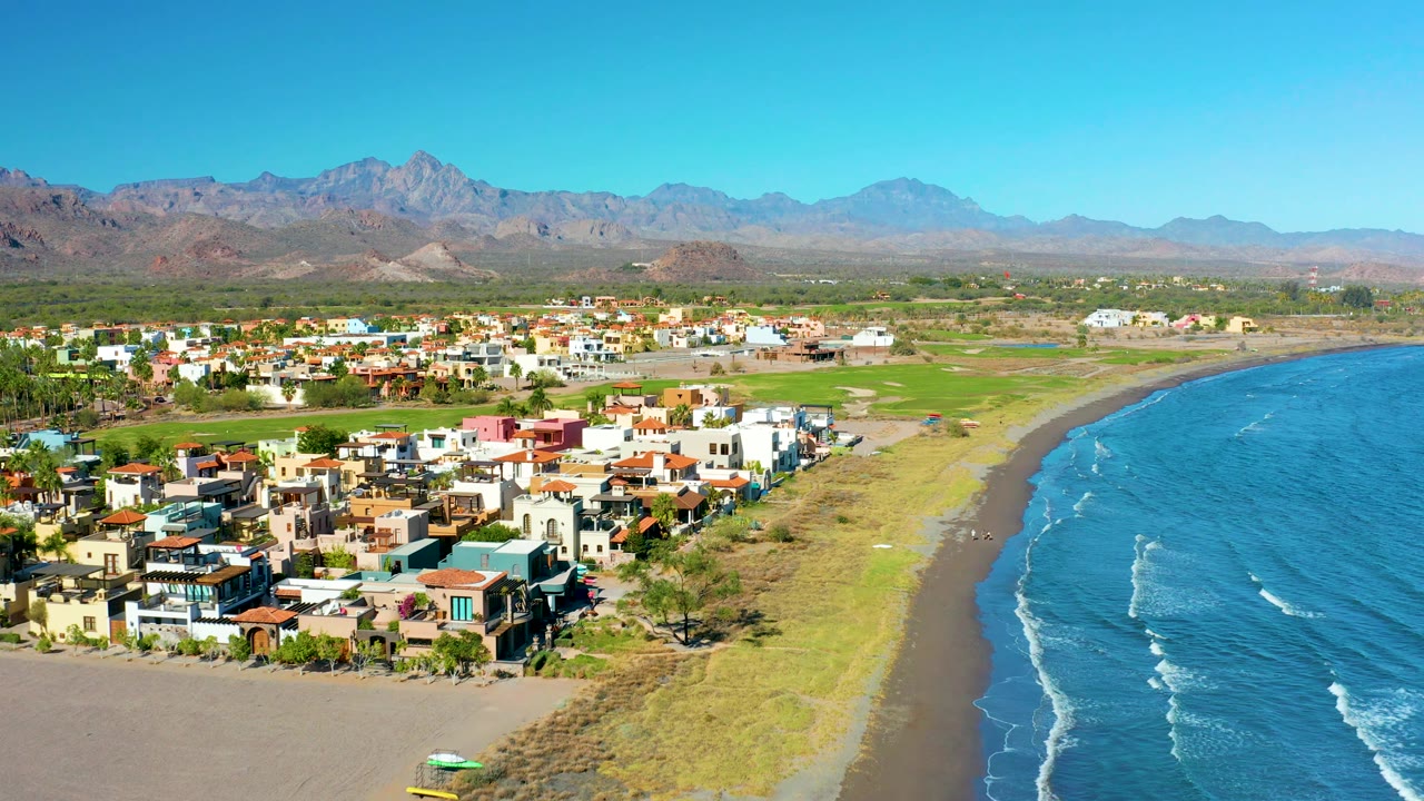Wide aerial frame over Loreto, Baja California Sur with more shoreline visible on the right, compact colorful residences on the left, green open land in the center, distant mountains, and no visible people.