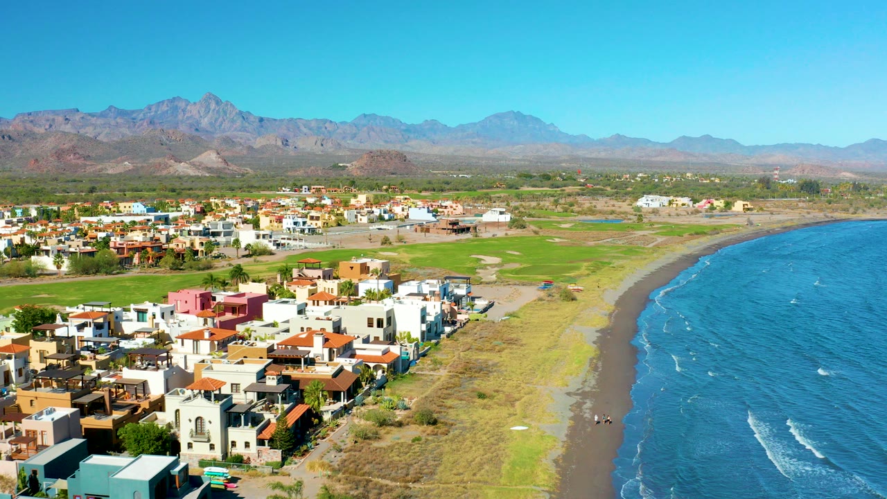 Aerial view over Loreto, Baja California Sur, with colorful homes in the foreground, a dark sand beach and blue ocean to the right, mountains in the distance, and no visible people.