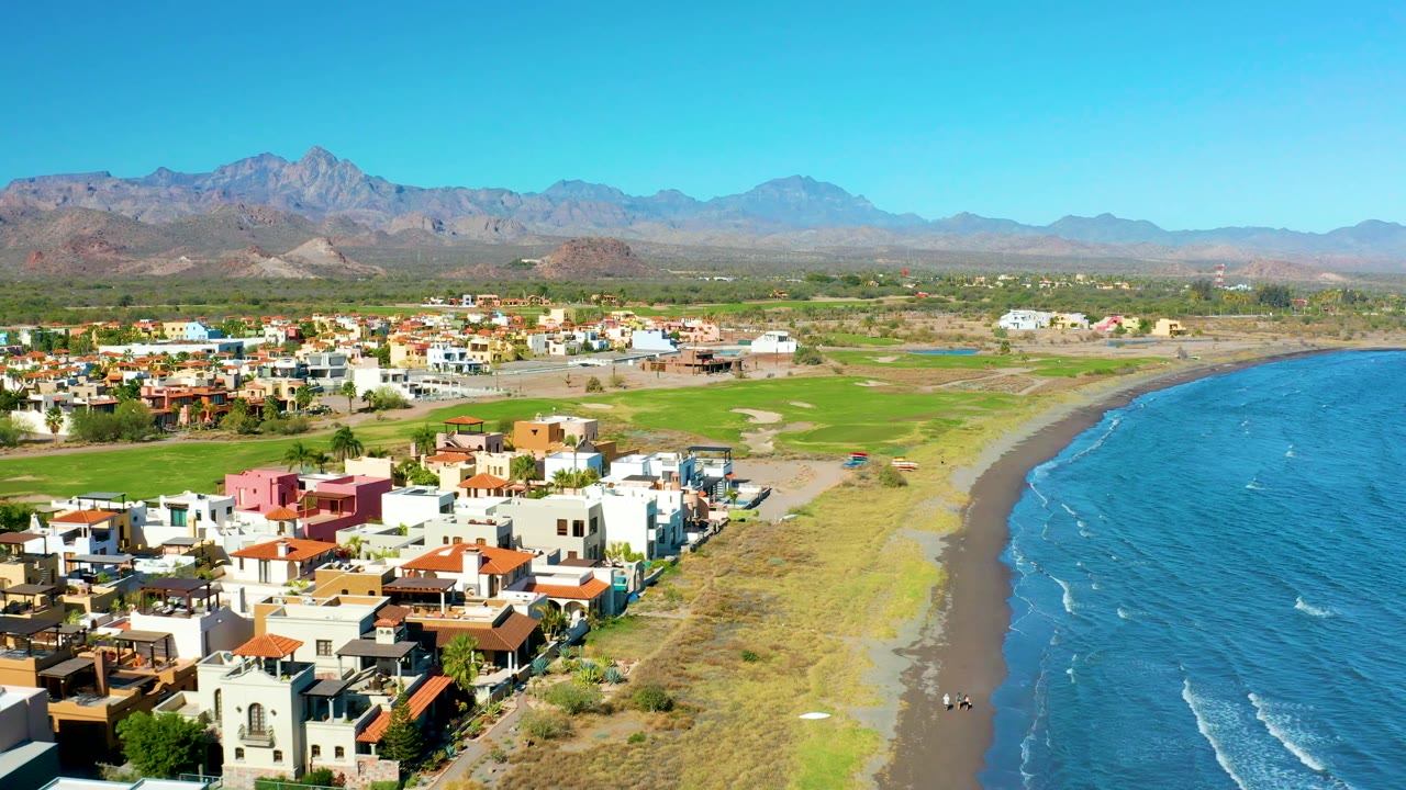 Aerial view in Loreto with homes lower left, beach grass and dark sand along the shore, small white wave lines on the ocean, distant mountains, and no visible people.