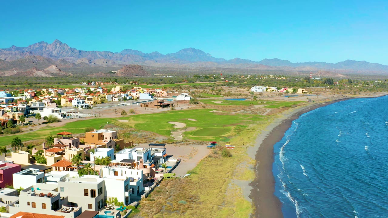 Drone view of beachfront homes beside a green field and curving shoreline.