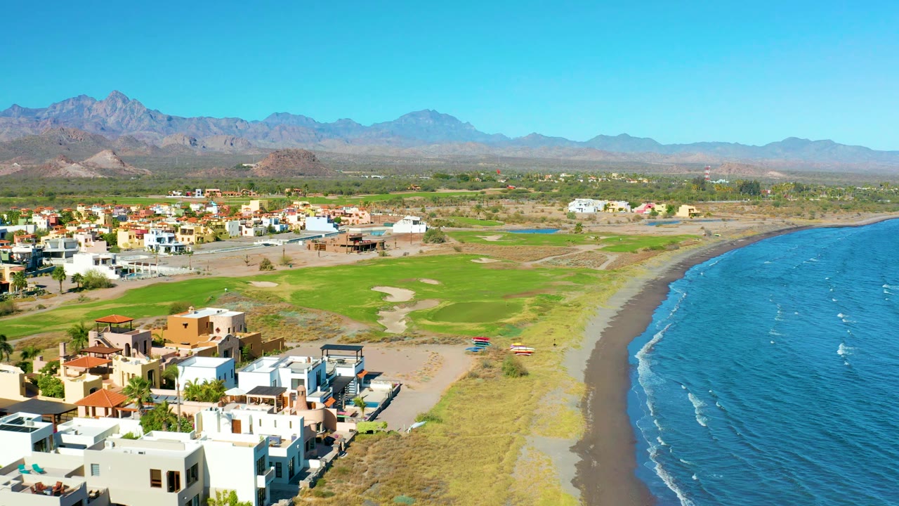 Wide aerial frame over Loreto showing residential blocks to the left, open green field in the center, sandy beach and rippled blue ocean to the right, hazy mountains beyond, and no visible people.