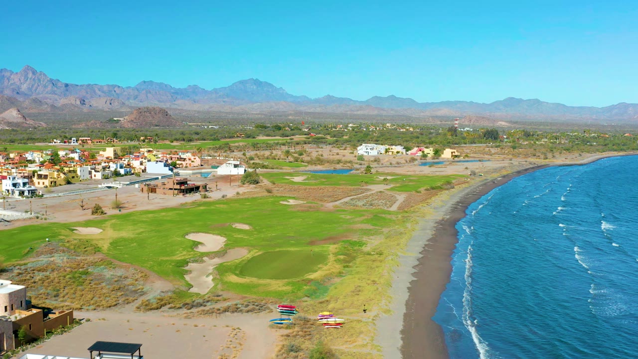 Aerial view in Loreto, Baja California Sur, showing the golf course closer to center, small colorful boats near the lower beach, blue water to the right, low buildings to the left, and no visible people.
