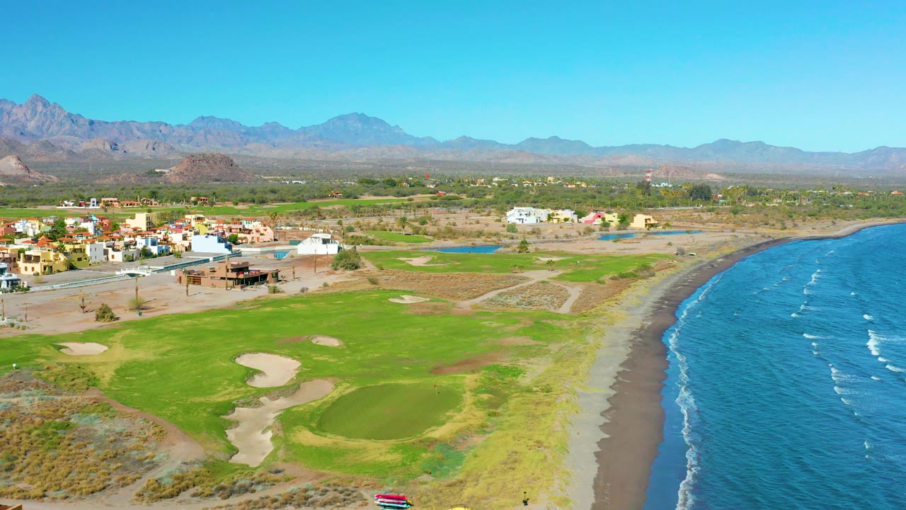 Golf course, beach, ocean, settlement, and distant mountains under clear sky.