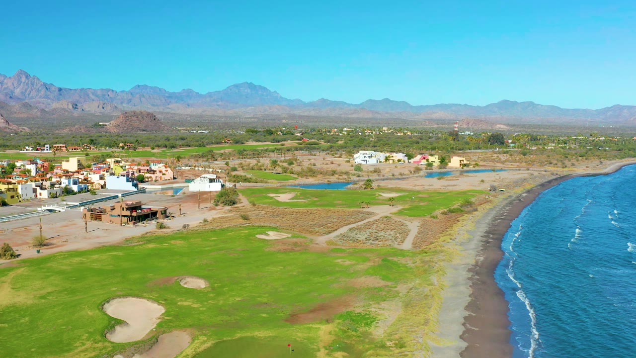 Aerial view in Loreto, Baja California Sur with bright green fairway in the foreground, sand traps, a curved beach and blue water to the right, homes to the left, and no visible people.