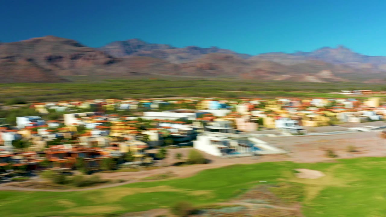 Motion-blurred aerial frame in Loreto, Baja California Sur showing residential buildings, green golf grass, sand traps, brown mountains, blue sky, and no visible people.