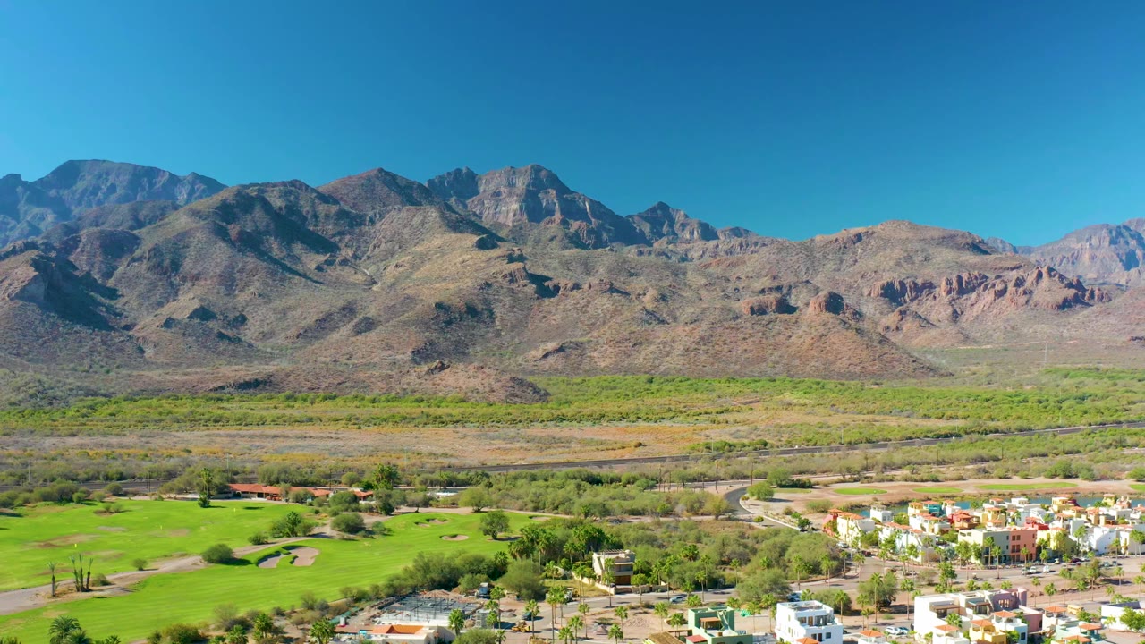 A wide aerial frame over Loreto, Baja California Sur, showing green fields, scattered residential buildings, a road, rugged mountains, clear blue sky, and no visible people.