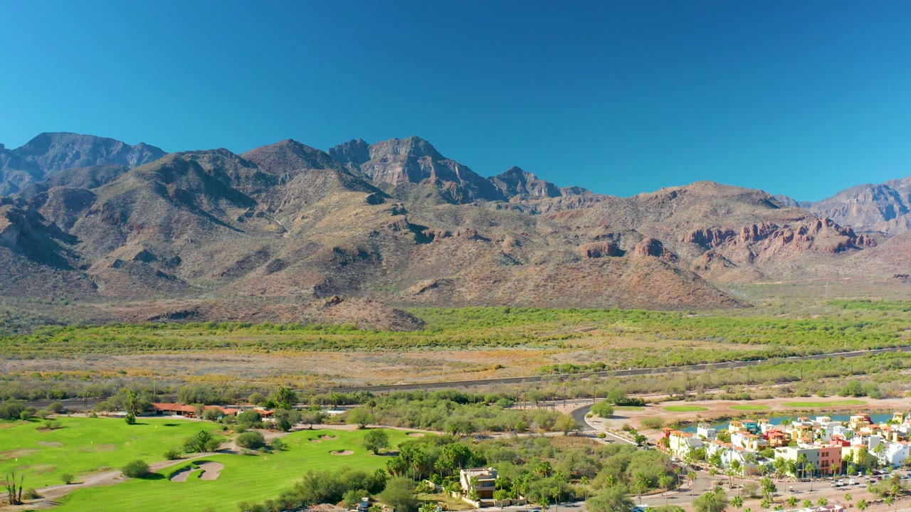 Wide aerial frame in Loreto, Baja California Sur showing rugged mountains, a clear blue sky, green fields, a road, and low residential buildings at the bottom, with no visible people.