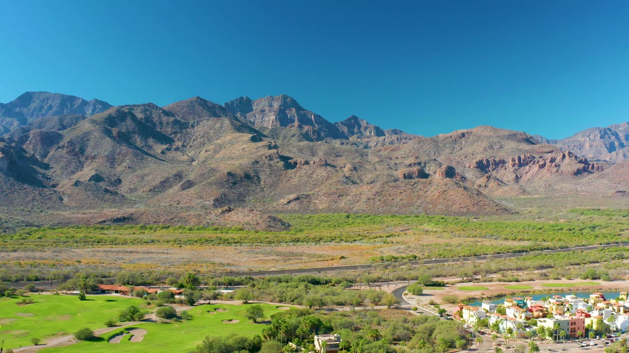 The frame shows Loreto, Baja California Sur mountains stretching across the horizon, with dry valley terrain, green ground, a roadway, and residential buildings near the bottom right, with no visible people.