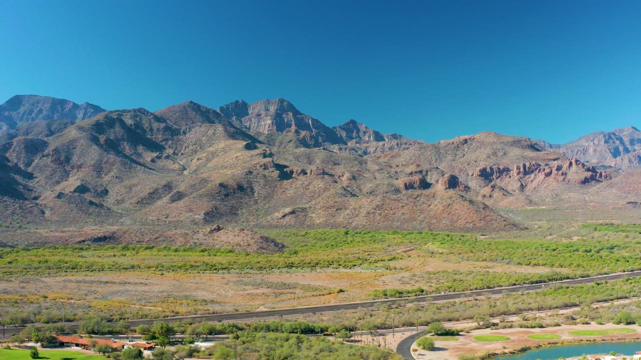 Opening aerial frame in Loreto, Baja California Sur, showing a paved road, green valley floor, distant rugged mountains, small buildings near the bottom edge, and no visible people.