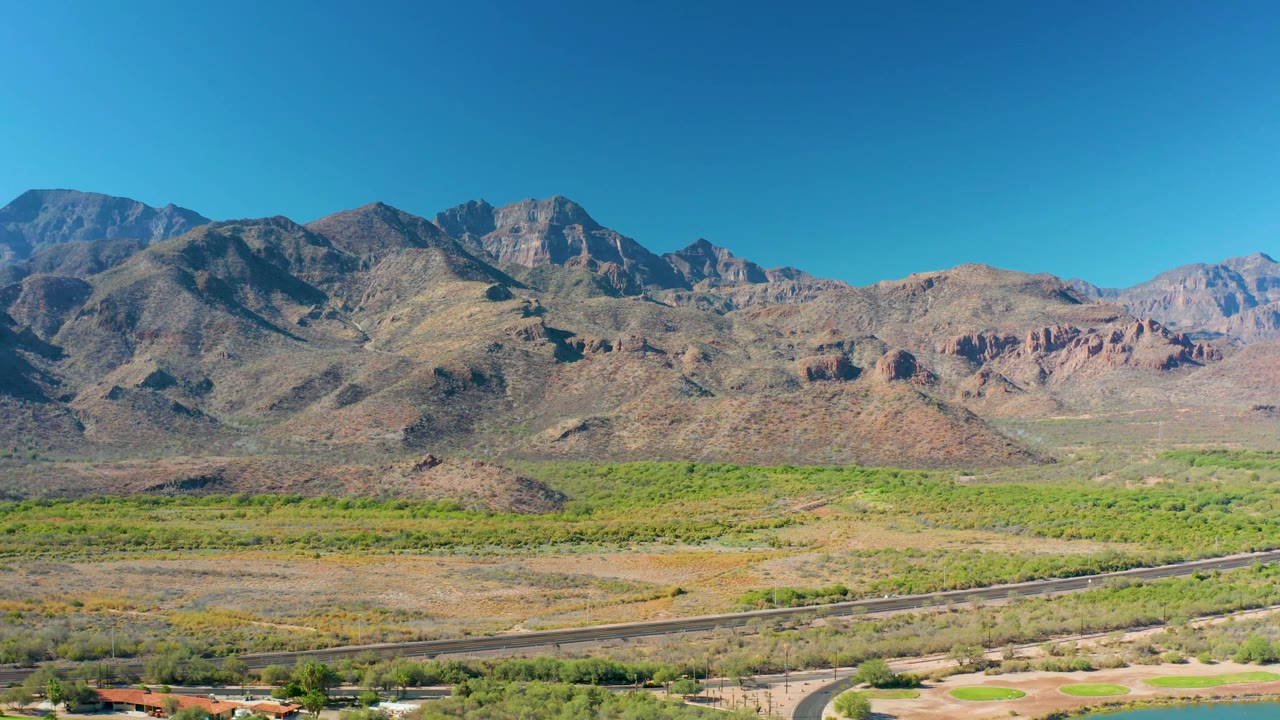Aerial frame in Loreto, Baja California Sur, showing the road low in frame, scrubland and green valley vegetation across the center, rocky peaks on the horizon, and no visible people.