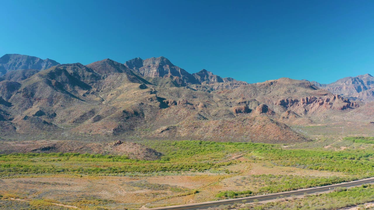 Early frame in Loreto, Baja California Sur showing the mountain range slightly farther across the valley, with green vegetation bands, dry brown ground, a curving road near the lower frame, and no visible people.