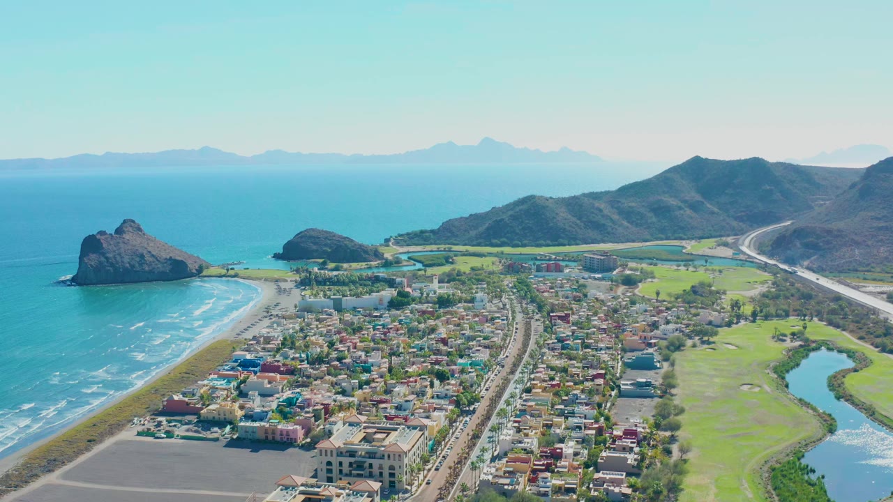Wide aerial view of shoreline, town blocks, fields, road, ocean rocks, and distant mountains.