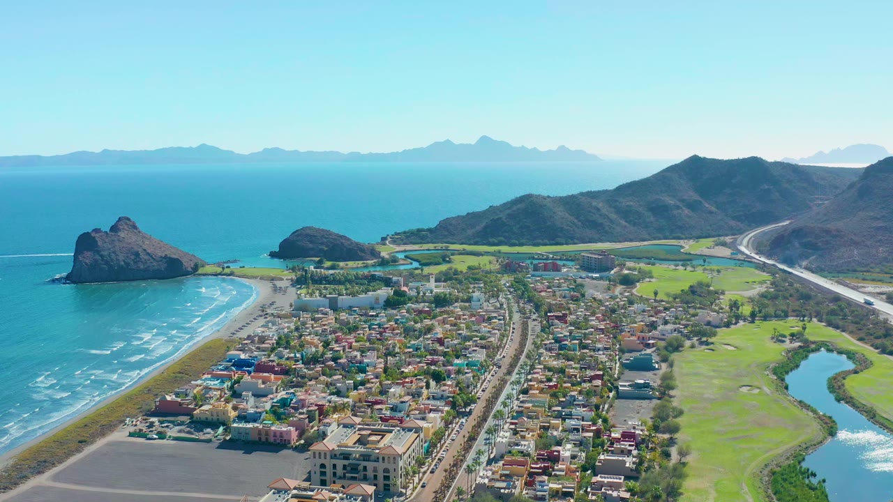 Opening aerial frame over Loreto, Baja California Sur, showing turquoise surf along a sandy beach, town blocks, a green river corridor, mountain ridges, distant islands, and no visible people.