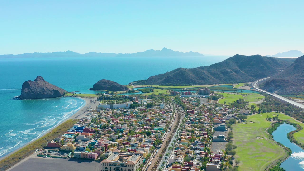 Aerial view over Loreto, Baja California Sur, with town blocks beside a sandy beach, a highway, blue ocean, dark hills, distant mountains, and no visible people.