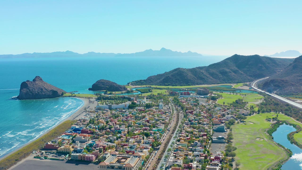 Aerial view over Loreto with the ocean and beach on the left, colorful low buildings in the lower frame, highway traffic through the town, green fields at right, and no visible people.