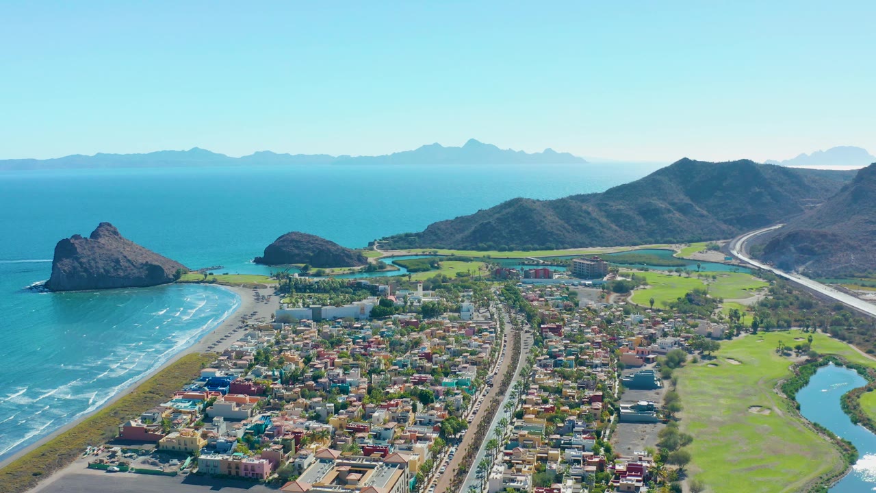 Wide aerial of ocean, rock formations, town, road, fields, and mountains.