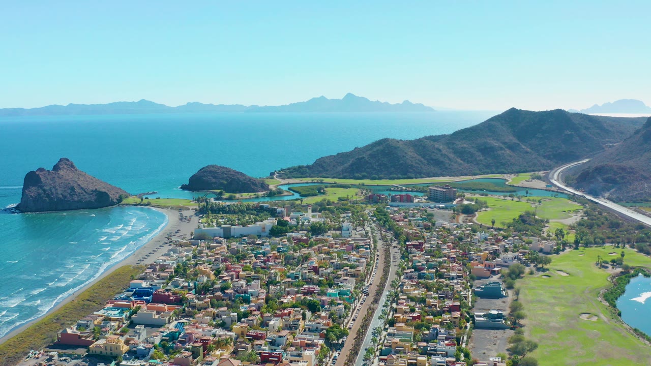 Opening aerial frame over Loreto, Baja California Sur, showing surf on a curved beach, a residential neighborhood, green golf course areas, a coastal road, dark hills, distant mountains, and no visible people.