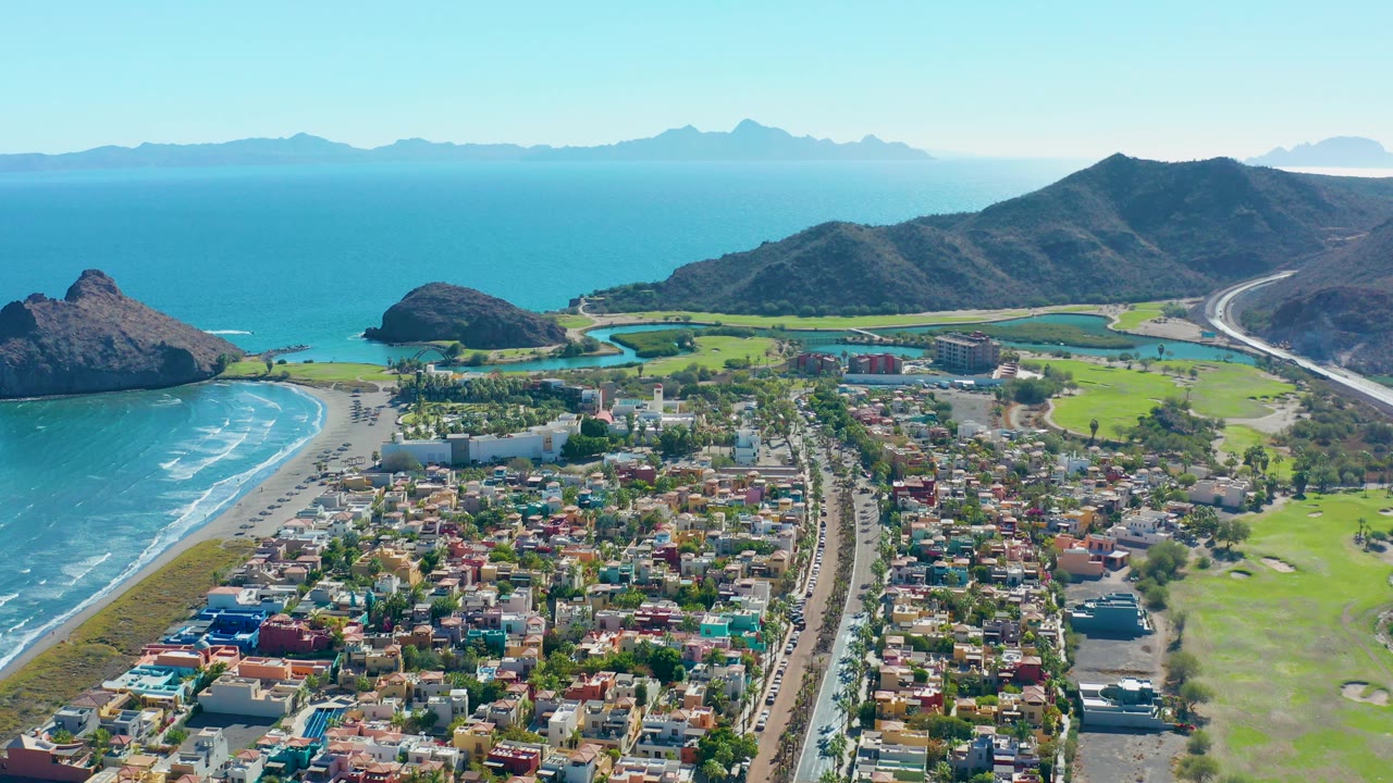Aerial frame over Loreto showing the beach curve and surf at left, dense low-rise buildings in the foreground, a central road, green fairways to the right, dark hills, distant blue mountains, and no visible people.