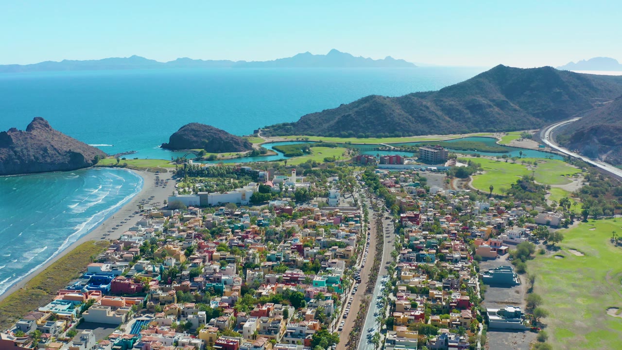 Wide aerial over Loreto with the shoreline and pale sand on the left, colorful roofs and streets below, golf course greens on the right, a rocky headland near the water, layered mountains on the horizon, and no visible people.