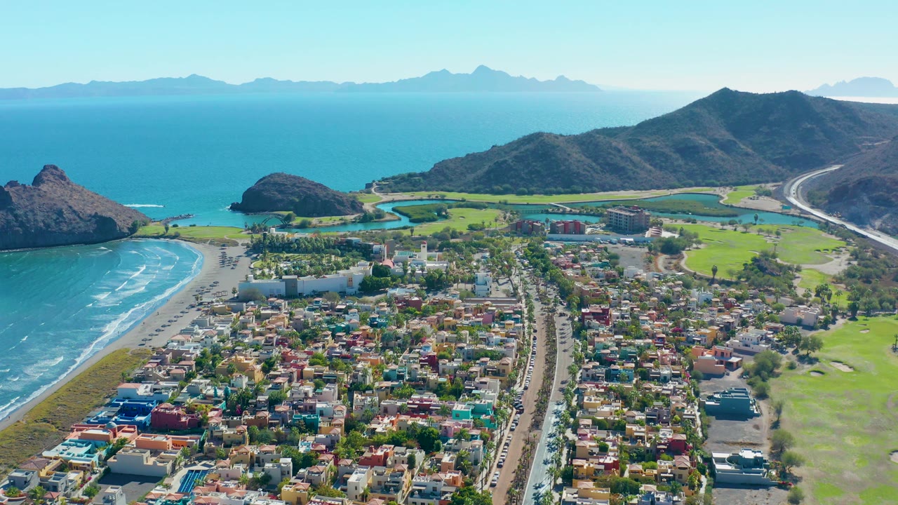 Aerial coastline view with town foreground, waves, green course, hills, and sea.