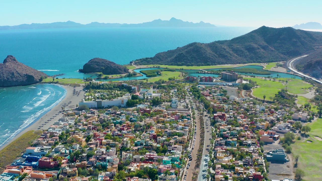 Aerial view over Loreto, Baja California Sur, with colorful homes in the foreground, a beach and golf greens near the shore, blue ocean beyond, mountains on both sides, and no visible people.