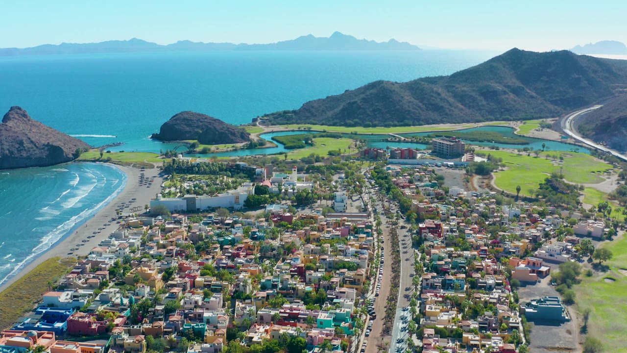 Aerial view across Loreto with the neighborhood filling the lower frame, the beach curving along the left, golf course greens and water features near the center, mountains behind, and no visible people.