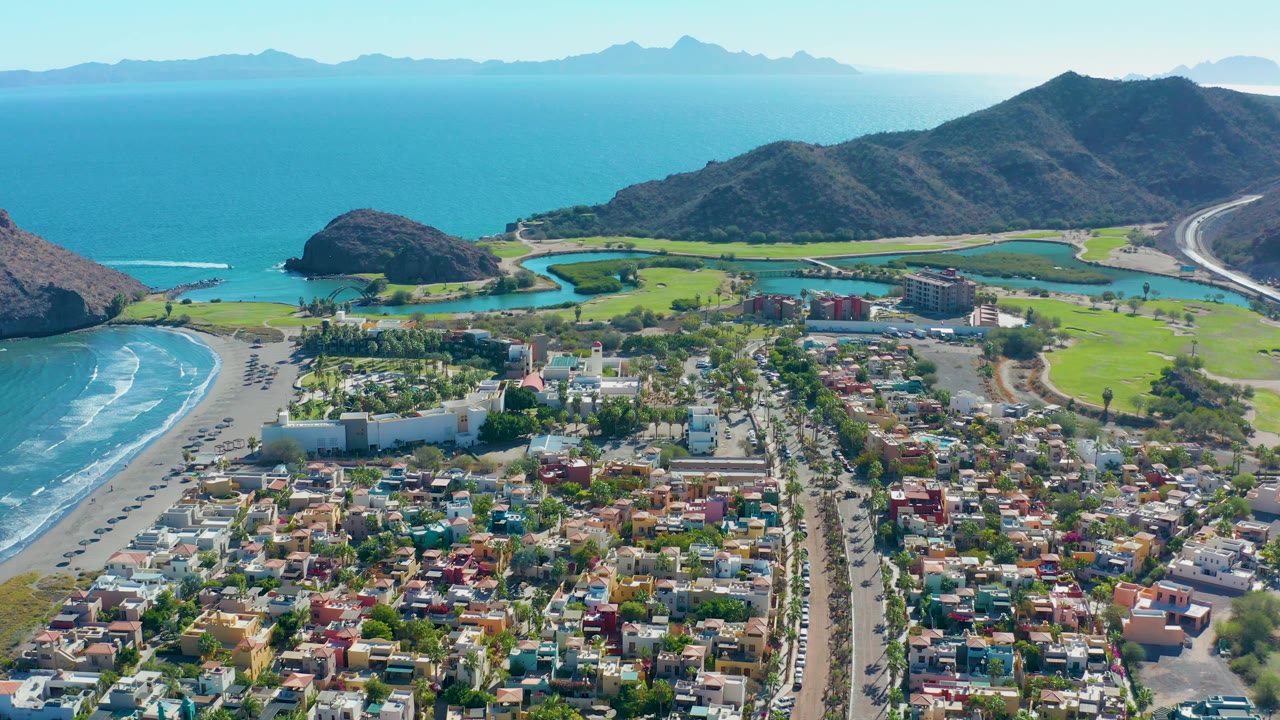 Aerial frame over Loreto showing a central road through low residential buildings, surf curling onto the left beach, bright green golf grounds near the lagoon-like channels, distant ocean islands, and no visible people.