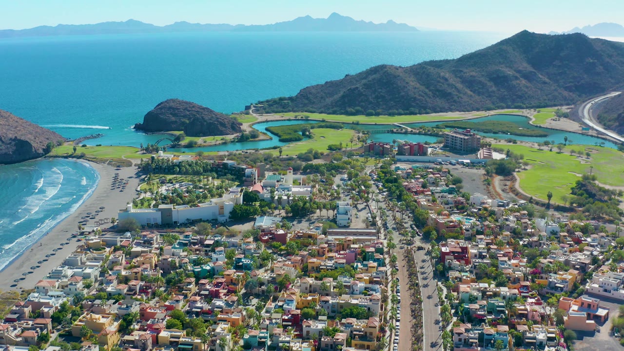 Drone aerial of Loreto beach town with green golf course and mountains along the coast.