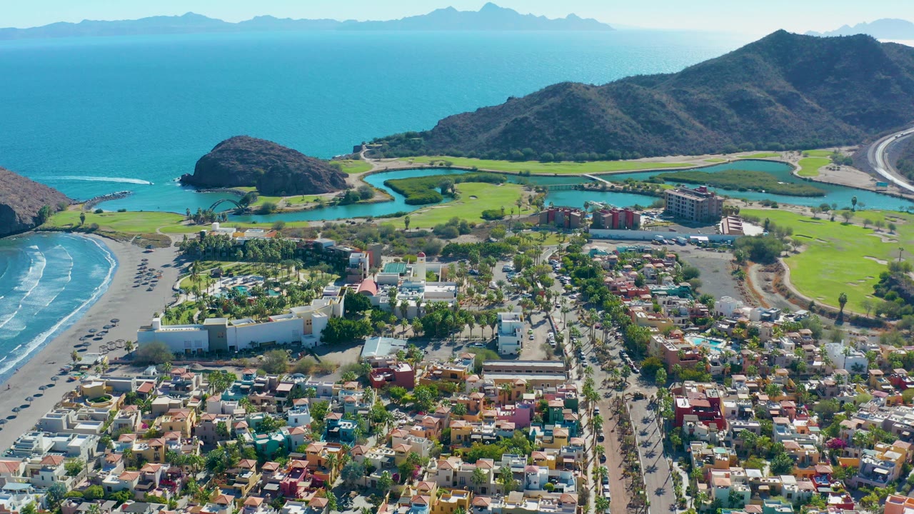 High aerial view over Loreto, Baja California Sur, showing a sandy beach at left, town buildings below, green golf fairways, blue ocean, hills, and no visible people.