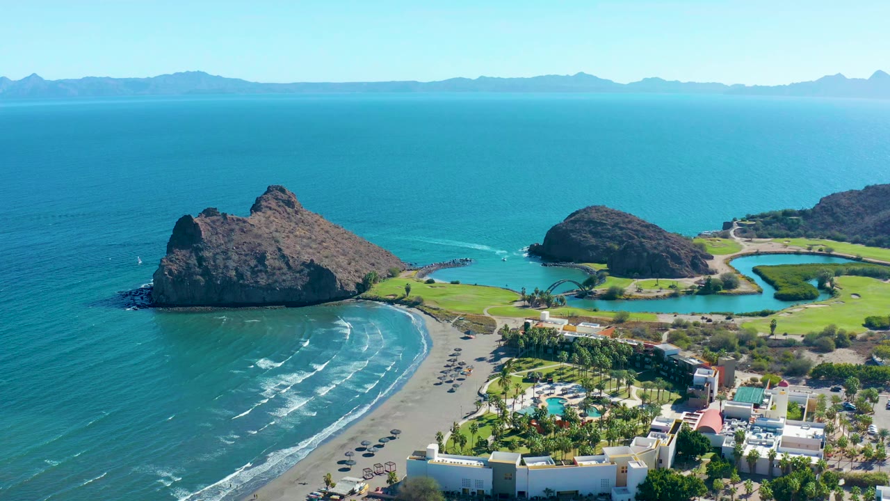 Aerial frame over Loreto showing the curved beach and surf slightly farther along the shoreline, with dark rocky outcrops, resort buildings, green grounds, distant mountains, and no visible people.