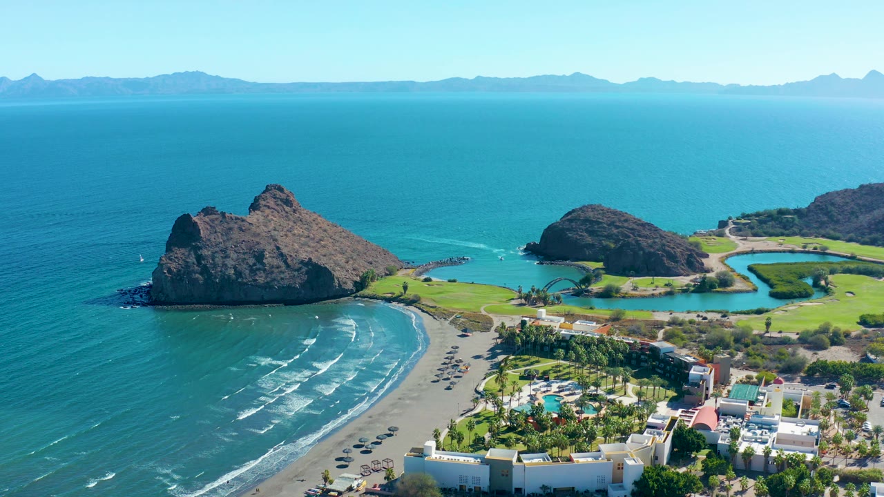 Mid-clip aerial frame over Loreto with more open water above the beach, dark rock formations on the shoreline, resort grounds in the lower right, distant mountain silhouettes, and no visible people.