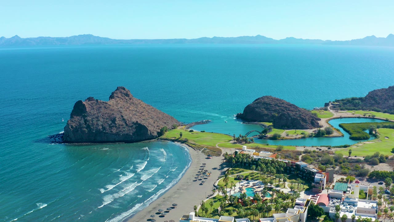 Aerial frame over Loreto, Baja California Sur, showing rocky outcrops in blue ocean, a curved beach with small waves, shoreline buildings, green grounds, distant mountains, and no visible people.