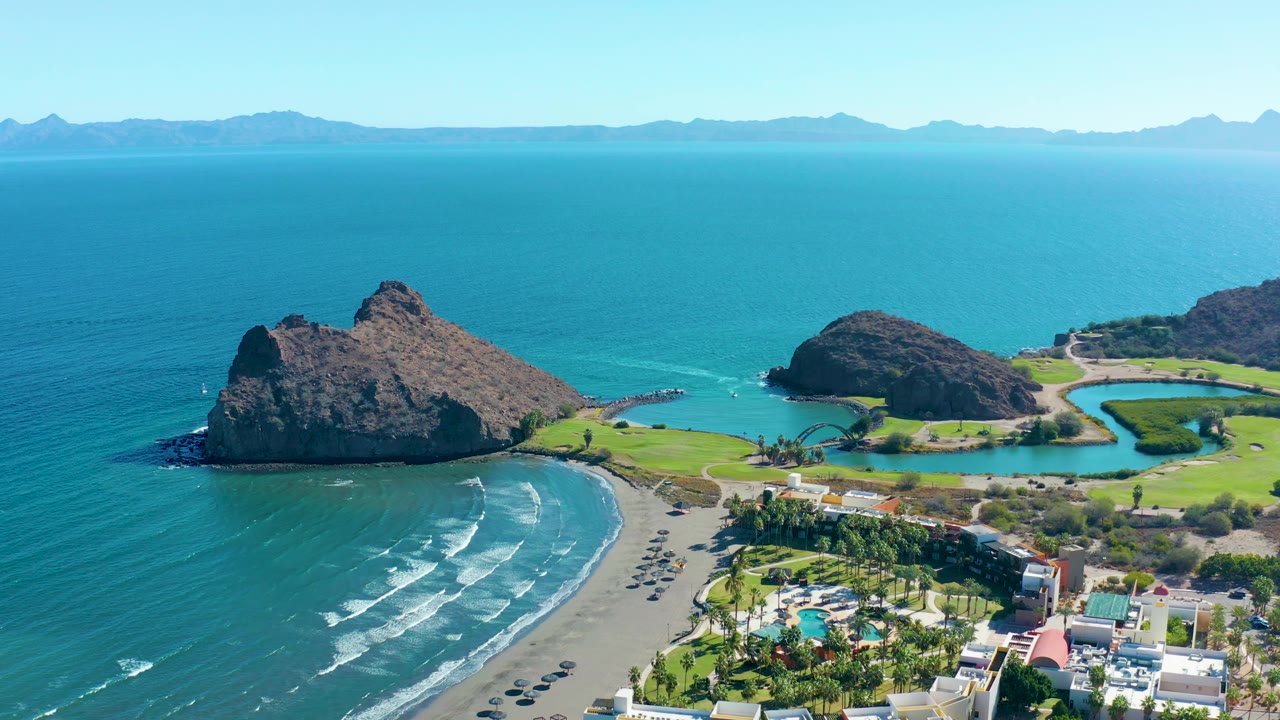 Aerial view in Loreto, Baja California Sur, with the large left rock slightly more centered, waves crossing the sandy beach, vehicles near the shore road, green areas, buildings, and no visible people.