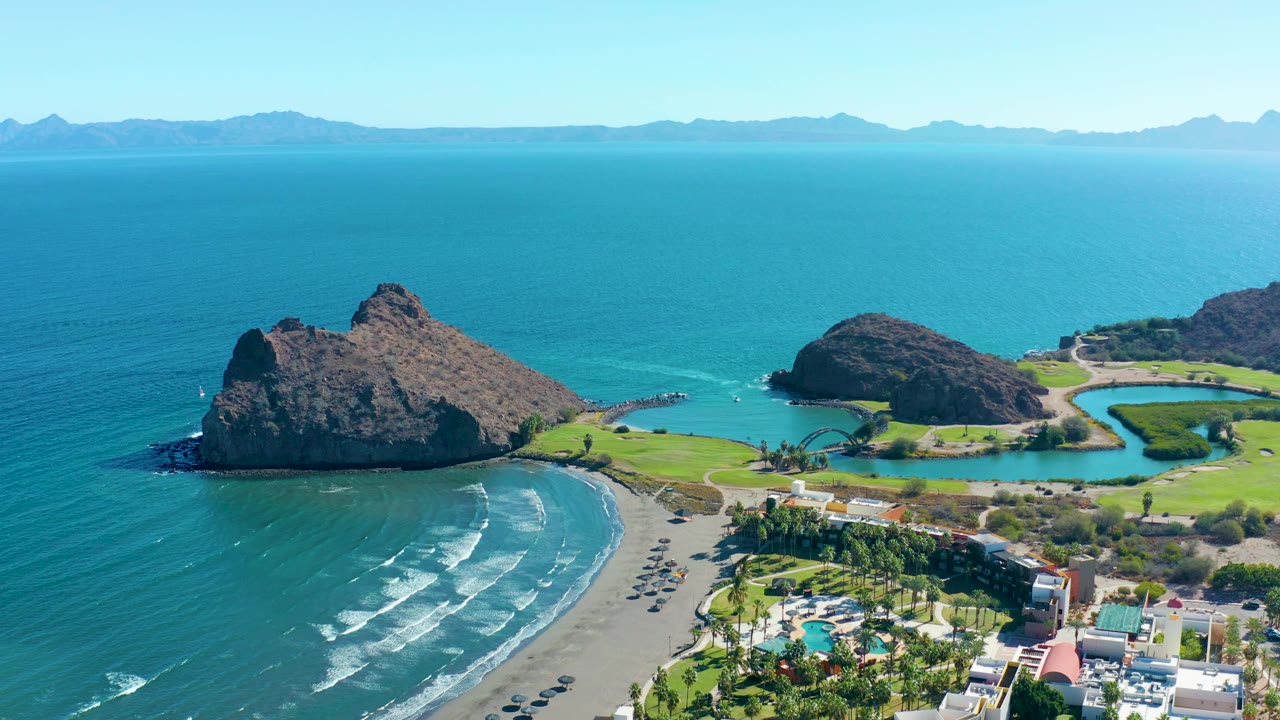 Aerial frame over Loreto, Baja California Sur, showing two rocky outcrops separated by turquoise water, a wave-lined beach, low shoreline buildings, green grounds, distant mountains, and no visible people.
