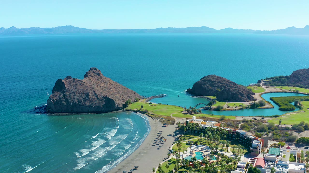 In Loreto, Baja California Sur, the aerial view shifts lower over the beach, with the large rock at left, surf bands on the sand, parked vehicles near the shore, green shoreline grounds, buildings, and no visible people.