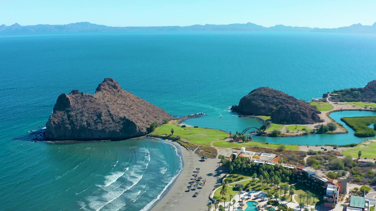 Drone-style aerial frame over Loreto showing the main rocky outcrop, a smaller outcrop to the right, crescent beach, turquoise water, shoreline buildings, and no visible people.