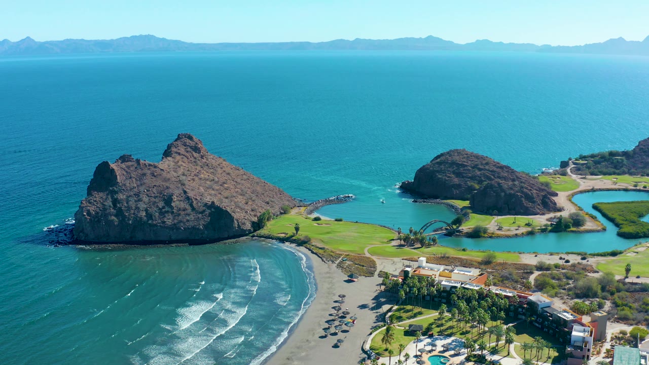 Aerial frame in Loreto with the rocky outcrop shifted farther left, a broad field of blue water, small waves reaching the sand, beachfront structures at lower right, and no visible people.