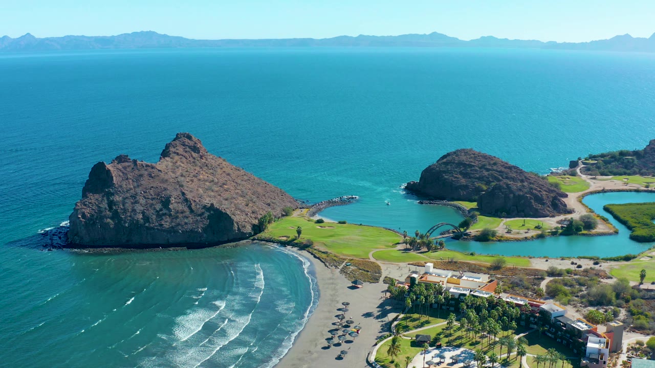 Loreto aerial view with the large rocky outcrop lower in frame, surf bands on the crescent beach, green lawn beside clear water, waterfront buildings, parked cars, and no visible people.