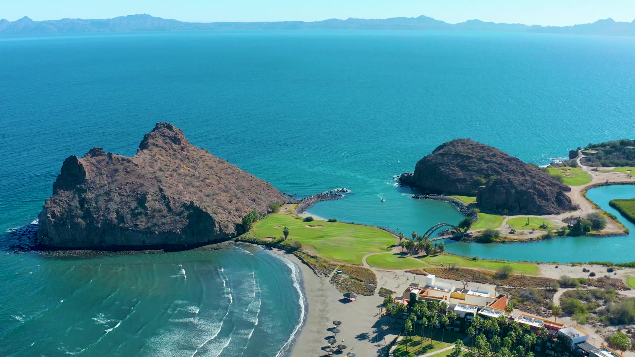 Opening aerial frame over Loreto, Baja California Sur, Mexico, showing two rocky islands, turquoise ocean, a curved beach, green grass, distant mountains, and no visible people.