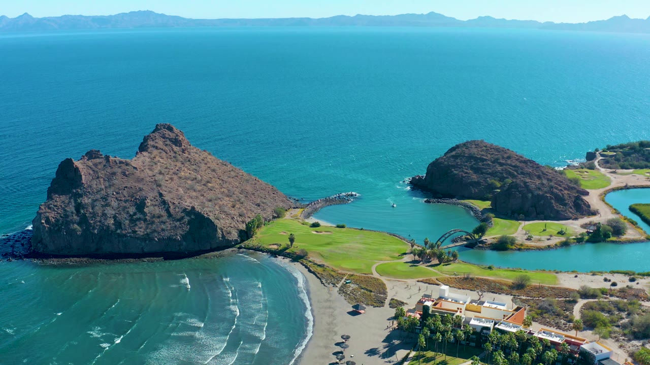 Aerial frame over Loreto, Baja California Sur, Mexico, showing the rocky island group, open turquoise water, beach surf, shoreline buildings, parked cars, distant mountain silhouettes, and no visible people.