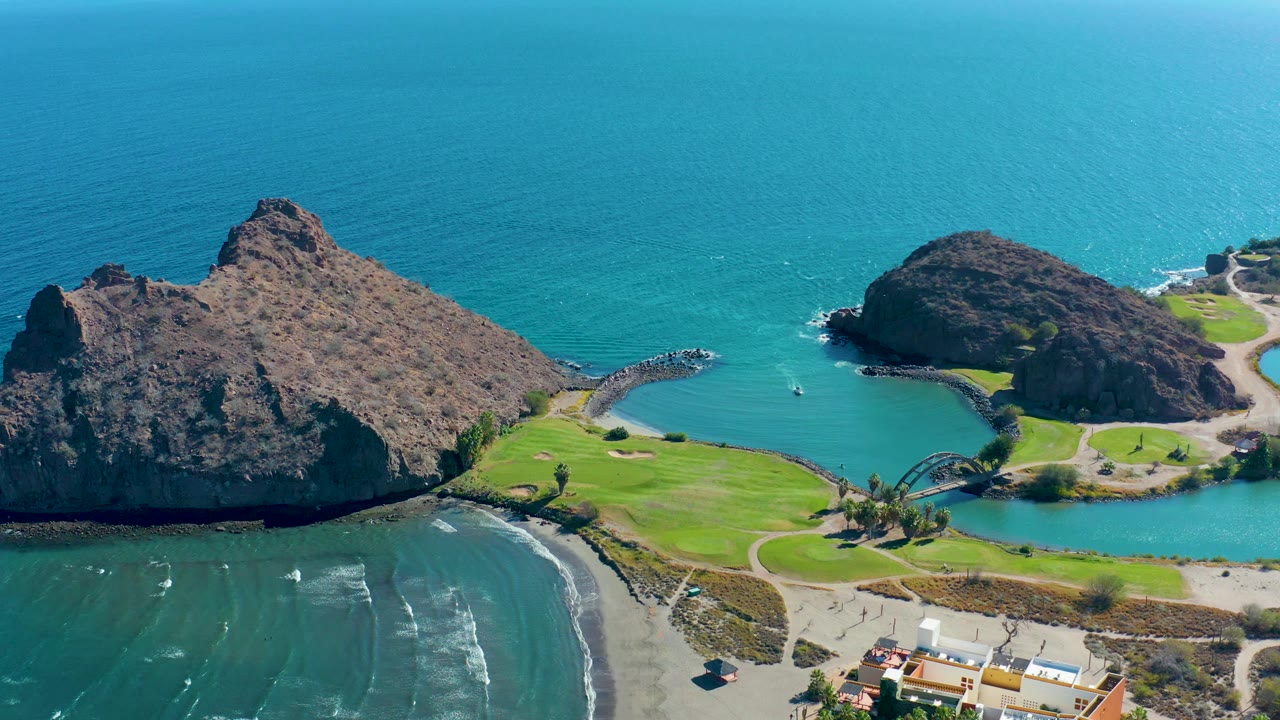Aerial frame over Loreto, Baja California Sur with turquoise cove water between two rocky headlands, bright grass near the shore, pale beach below, a small boat in the water, and no visible people.