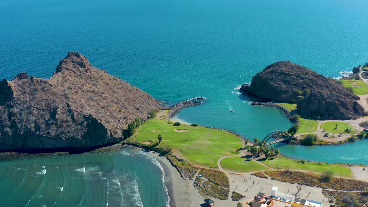 Aerial frame over Loreto, Baja California Sur with a large rocky point on the left, a rounded headland on the right, blue cove water, green grass, beach, buildings near the bottom, and no visible people.