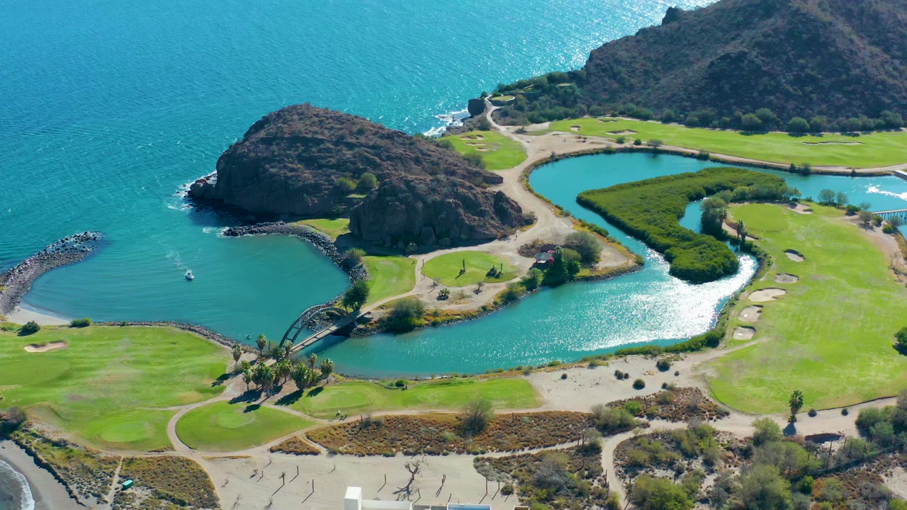 Aerial frame over Loreto, Baja California Sur showing turquoise water, a curving inlet, green fairways, rocky outcrops, and no visible people at the start of the clip.
