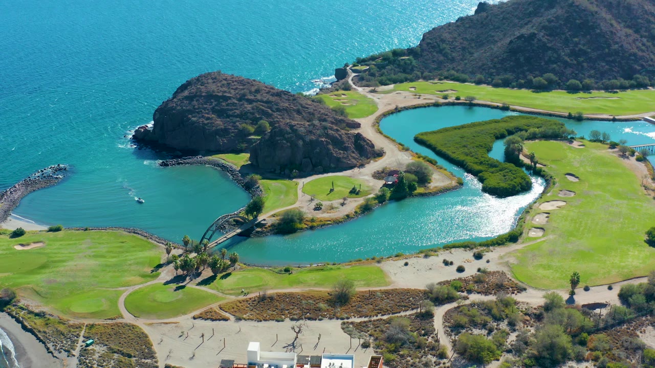 Aerial view in Loreto, Baja California Sur with the lagoon reflecting sunlight, rocky hills behind it, green grounds to the right, a bridge near the shore, and no visible people.